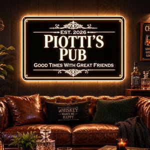 May include: A lit sign that reads "PIOTTI'S PUB" above a brown leather couch. The sign also says "EST. 2026" and "GOOD TIMES WITH GREAT FRIENDS". A small sign says "CHEERS" with glasses clinking. A pillow says "WHISKEY MAKES ME HAPPY".