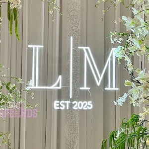 May include: A neon sign displaying the letters "LM" with "EST 2025" underneath, surrounded by white and cream-colored flowers and greenery. The sign is centered against a backdrop of white curtains.