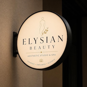 Peut inclure: Panneau rond et lumineux pour "ELYSIAN BEAUTY", studio et spa esthétique. Le panneau présente un cadre noir, une face crème avec des lettres dorées et un dessin au trait d'une silhouette et de feuilles. Le panneau comprend également les mots "Enhance, Empower, Elevate."