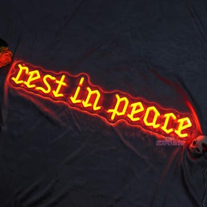 K&ouml;nnte beinhalten: Ein Neon-Schild mit den Worten "rest in peace" in einer gotischen Schriftart. Das Neon ist gelb und der Hintergrund ist schwarz.