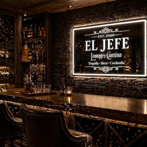 Puede incluir: Interior de bar oscuro con un letrero luminoso "EL JEFE Loungey Cantina". El texto "Tequila, Beer, Cocktails" es visible. El bar tiene una encimera de madera, sillas de cuero y estantes llenos de botellas.