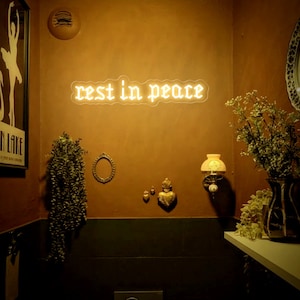K&ouml;nnte beinhalten: Ein wei&szlig;es Neon-Schild mit den Worten "rest in peace" in gotischer Schrift, das an einer braunen Wand h&auml;ngt. Das Schild wird mit einem warmen gelben Licht beleuchtet. An der Wand befinden sich mehrere Dekorationsartikel, darunter ein kleiner Spiegel, eine Vase mit getrockneten Blumen und eine kleine goldene Statue.