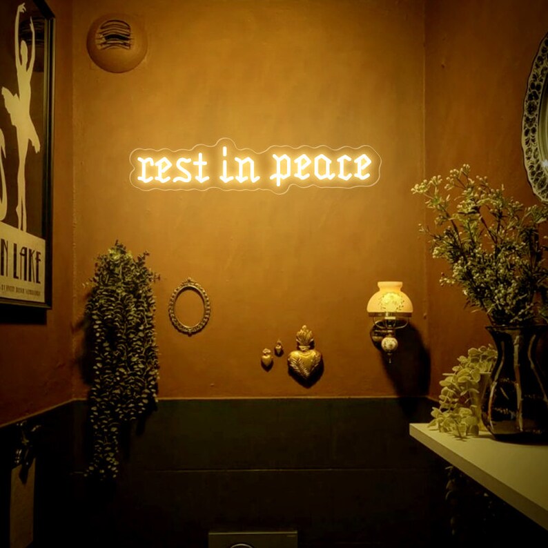 K&ouml;nnte beinhalten: Ein wei&szlig;es Neon-Schild mit den Worten "rest in peace" in gotischer Schrift, das an einer braunen Wand h&auml;ngt. Das Schild wird mit einem warmen gelben Licht beleuchtet. An der Wand befinden sich mehrere Dekorationsartikel, darunter ein kleiner Spiegel, eine Vase mit getrockneten Blumen und eine kleine goldene Statue.