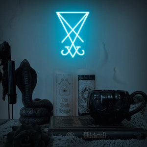 May include: A blue neon sign depicting the sigil of Baphomet, a symbol associated with occultism and witchcraft. The sign is displayed on a wall with a black candle holder, a black snake statue, and a book titled "Witchcraft" in the foreground.