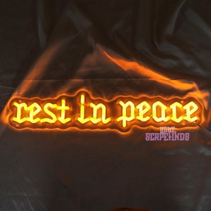 K&ouml;nnte beinhalten: Ein Neon-Schild mit den Worten "rest in peace" in einer gotischen Schriftart. Das Schild leuchtet in einem warmen Gelbton.