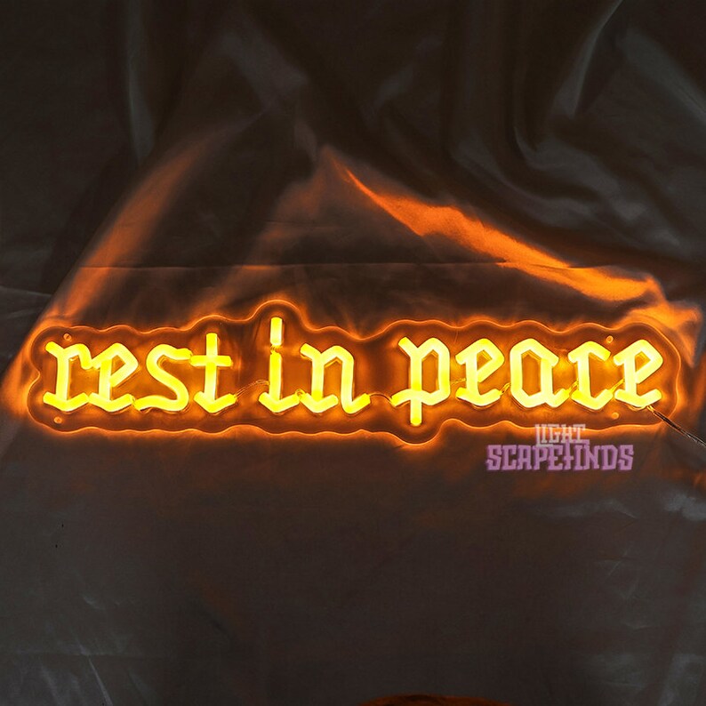 K&ouml;nnte beinhalten: Ein Neon-Schild mit den Worten "rest in peace" in einer gotischen Schriftart. Das Schild leuchtet in einem warmen Gelbton.