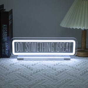 Puede incluir: Un letrero LED rectangular de acrílico transparente con las palabras "DO SOMETHING GREAT" en letras blancas iluminadas. El letrero está sobre una base transparente y se encuentra sobre una superficie blanca texturizada. Una lámpara y libros están en el fondo.