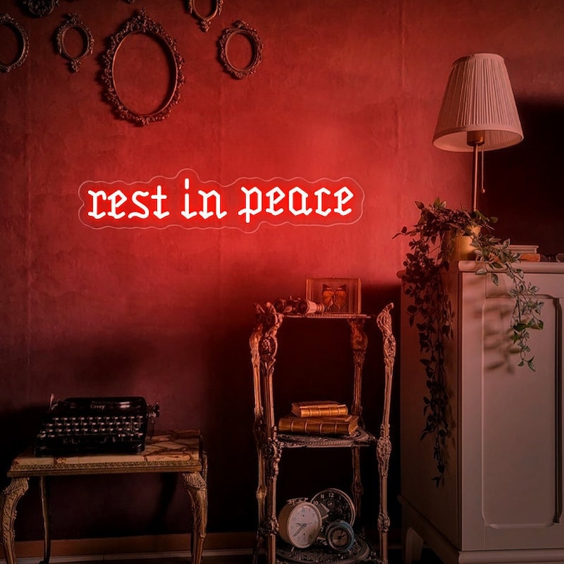 K&ouml;nnte beinhalten: Eine rote Wand mit einem wei&szlig;en Neon-Schild, auf dem in gotischer Schrift "rest in peace" steht. An der Wand befinden sich drei verzierte goldene Bilderrahmen, ein kleiner Beistelltisch mit B&uuml;chern und einer Uhr sowie ein hoher Schrank mit einer Pflanze darauf.