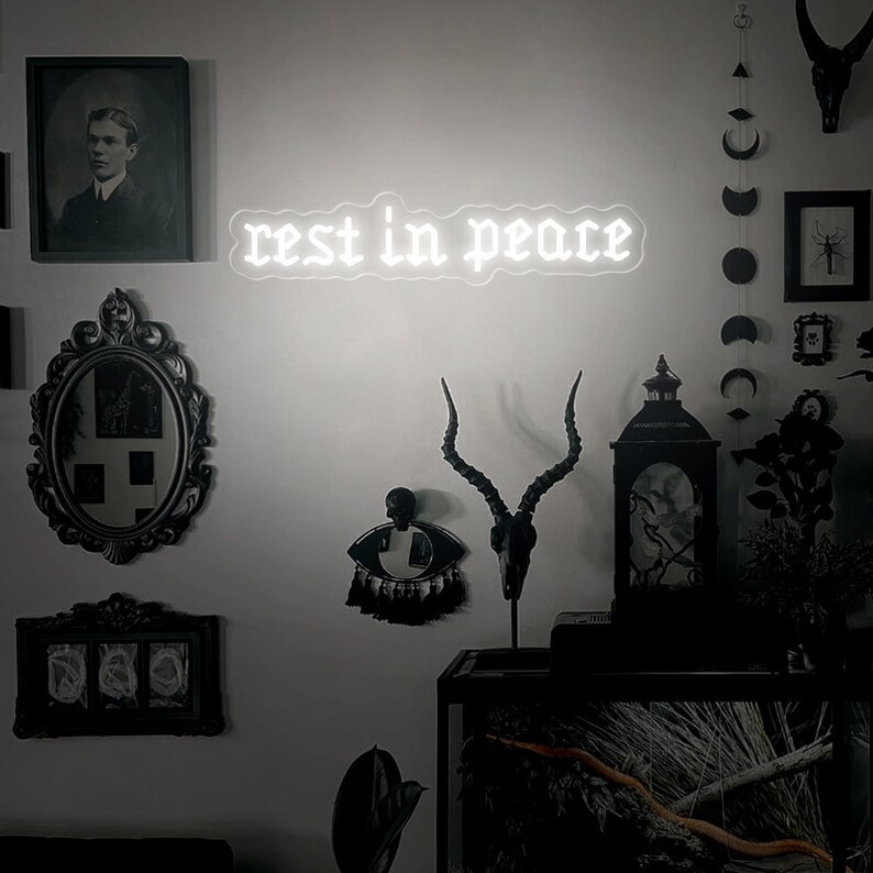 K&ouml;nnte beinhalten: Ein wei&szlig;es Neon-Schild mit den Worten "rest in peace" in gotischer Schrift. Das Schild h&auml;ngt an einer wei&szlig;en Wand mit schwarzer Dekoration, darunter ein Sch&auml;del mit Geweih, eine schwarze Laterne und gerahmte Bilder.