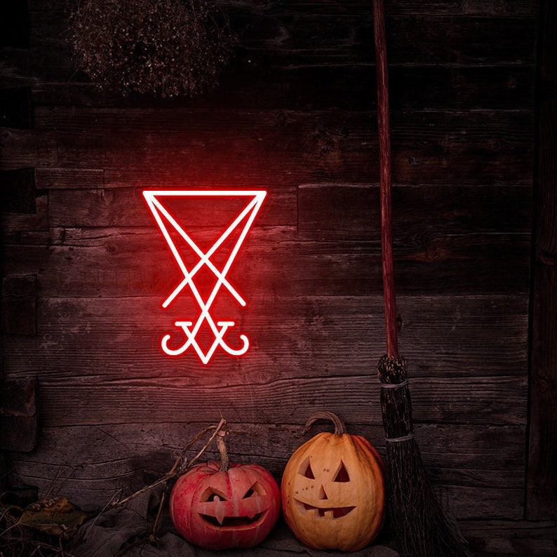 May include: A red neon sign depicting the sigil of Satan, a pentagram with an inverted cross, on a dark wooden wall. Two carved pumpkins sit in front of the sign, one with a traditional jack-o'-lantern face and the other with a more sinister expression. A broom leans against the wall.