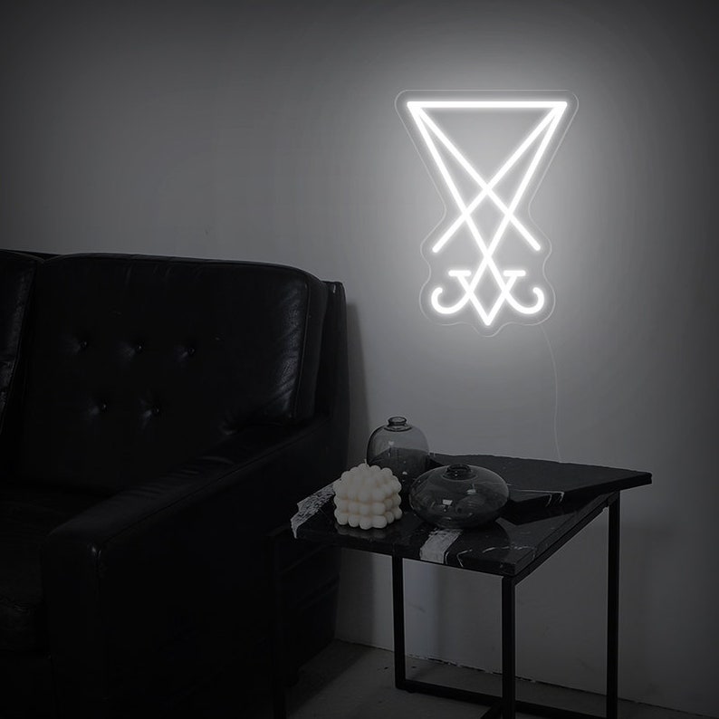 May include: White neon sign depicting the sigil of Baphomet, a symbol associated with Satanism, against a grey wall. The sign is mounted above a black leather sofa and a black side table with a white candle and a glass vase.