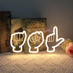 May include: White neon sign with three hand gestures in the shape of the letters "I Love You" in American Sign Language. The sign is illuminated and is sitting on a table with a book and a flower in the background.