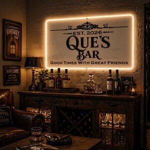 May include: A dimly lit bar scene featuring a sign that reads "QUE'S BAR" with the text "Good Times With Great Friends." The bar has a wooden cabinet with bottles, glasses, and a lamp. A leather armchair and a table with a glass of whiskey are in the foreground.
