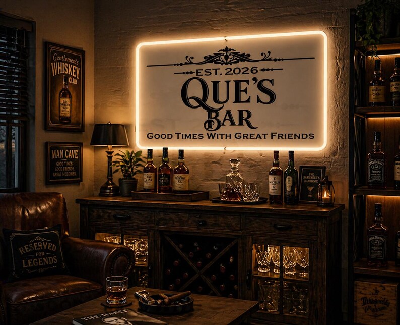 May include: A dimly lit bar scene featuring a sign that reads "QUE'S BAR" with the text "Good Times With Great Friends." The bar has a wooden cabinet with bottles, glasses, and a lamp. A leather armchair and a table with a glass of whiskey are in the foreground.