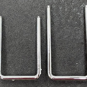 May include: Two chrome-plated, U-shaped metal objects. The objects are of different sizes, with rounded tops and bottoms. The metal has a shiny, reflective surface. The objects are set against a dark, textured background.