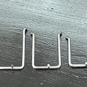 Op de afbeelding: Drie zilverkleurige, U-vormige metalen clips met een klein, rond detail aan de onderkant van elk. De clips zijn in een rij gerangschikt, waarbij de rechter clip de kleinste is.