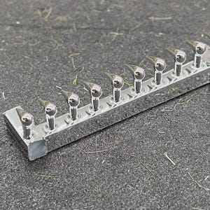 May include: A row of ten small, shiny, silver-colored metal objects. Each object has a rounded top and a pointed, triangular projection. They are mounted on a rectangular, silver-colored base. The objects are arranged in a straight line.