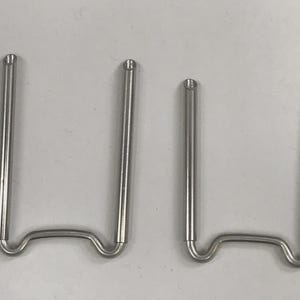 Puede incluir: Dos objetos metálicos plateados en forma de U. Cada uno tiene dos patas rectas y paralelas conectadas por una base curva. La parte superior de cada pata tiene una pequeña punta redondeada. Probablemente hardware o herramientas.