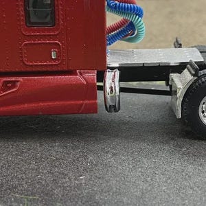 May include: A detailed side view of a red semi-truck model. The truck features a chrome wheel, black tyres, and a silver fender. The cab has a window and a door. The truck has blue, red, and white air lines.