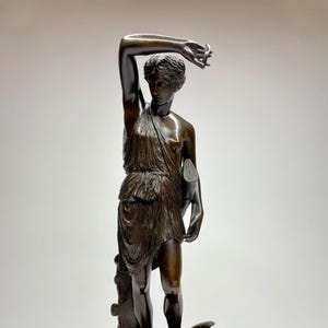 Puede incluir: Una escultura de bronce de una mujer de pie con el brazo levantado, sosteniendo un pequeño objeto en la mano. Lleva una túnica fluida y está de pie sobre una base con un diseño decorativo. La escultura está sobre una base de mármol negro.