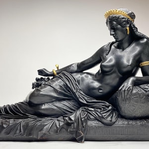 Puede incluir: Una estatua reclinada de una mujer con una corona y joyas, de color negro y dorado. La estatua está hecha de un material liso y pulido y descansa sobre una base negra.