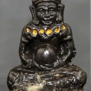 Puede incluir: Una figura oscura, sentada, con un sombrero alto y puntiagudo y una expresión serena. La figura tiene detalles dorados y está sentada sobre una base de calaveras. La estatua probablemente está hecha de un material oscuro, posiblemente metálico.