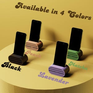 May include: Four smartphone stands in black, peach, lavender, and matcha green. Each stand holds a black smartphone and has a rounded base with horizontal vent-like detailing. The text "Available in 4 Colors" is at the top.
