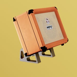May include: An orange amplifier with black corner protectors and the brand name "Orange" on the front. The amplifier is mounted on a gray stand. The speaker has a beige mesh cover.