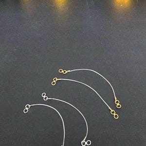 Può includere: Tre cordini elastici trasparenti con chiusure in metallo dorate e argentate. I cordini sono disposti a semicerchio su una superficie nera.