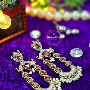 May include: A pair of silver and gold earrings with pink gemstones and pearl accents. The earrings feature a peacock design and are hanging from a purple velvet background.
