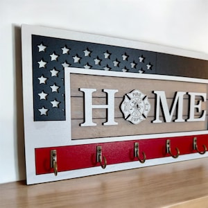 May include: Wooden key holder with a red, white, and blue American flag design. The word "HOME" is displayed in white letters with a firefighter's emblem in the center. Five hooks are attached to the bottom of the holder.