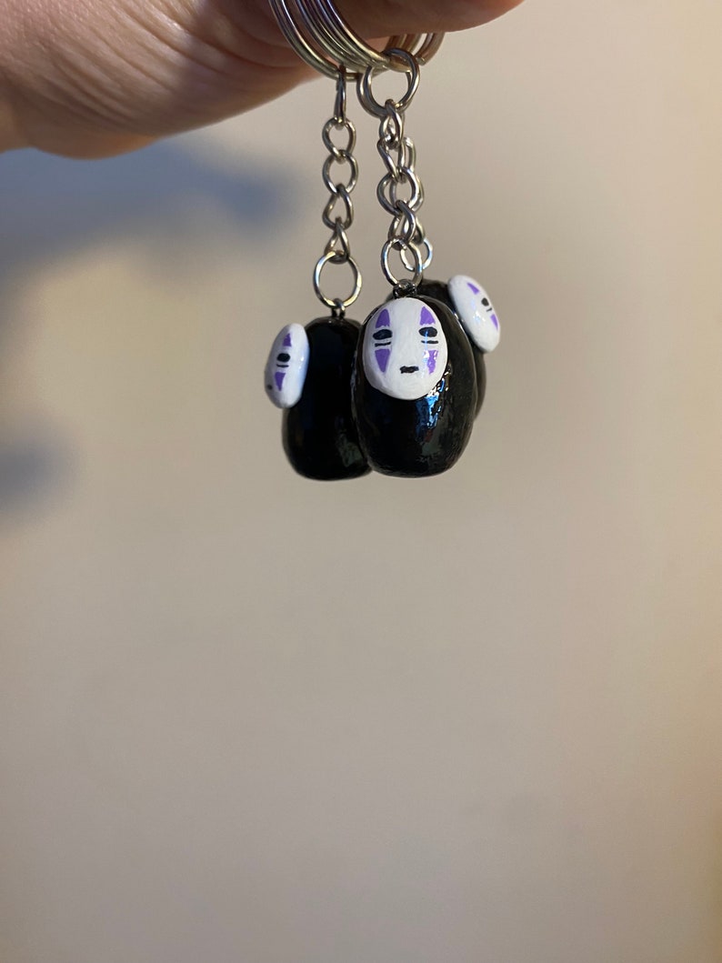 Puede incluir: Tres llaveros negros y blancos de No Face con detalles morados. Los llaveros est&aacute;n unidos a cadenas plateadas.