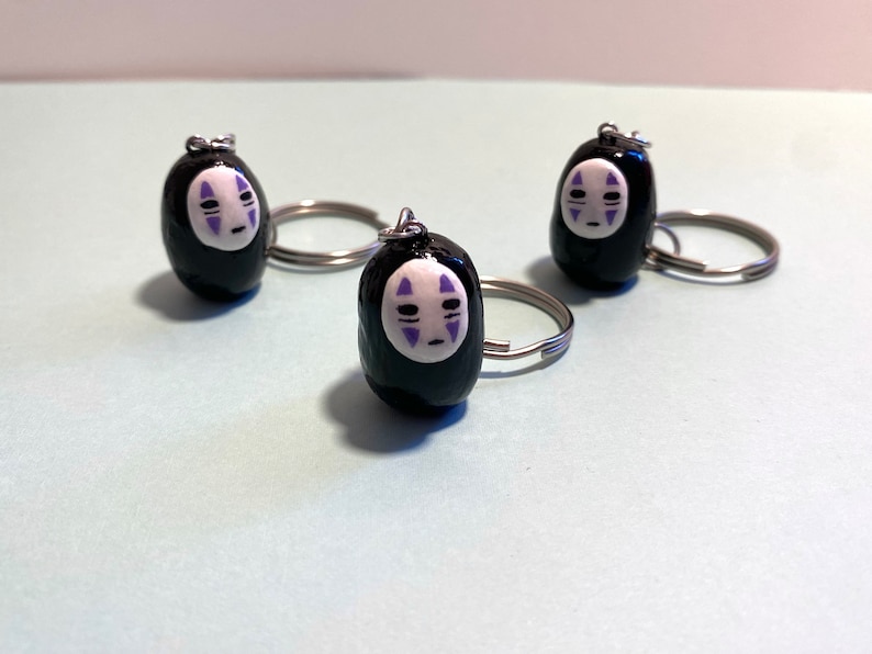 Puede incluir: Tres llaveros negros con anillas de plata. Cada llavero presenta una cara blanca con detalles morados, que se asemeja al personaje Sin Cara de la pel&iacute;cula animada El viaje de Chihiro.