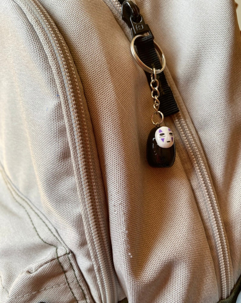 Puede incluir: Un llavero negro y blanco de No Face con detalles morados colgando de una cadena plateada en una bolsa gris con cremallera.
