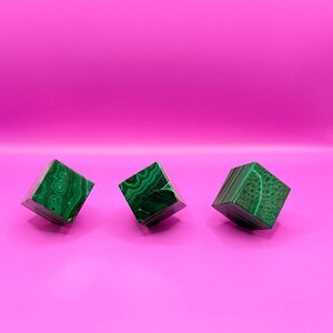 Può includere: Tre cubi di malachite verde con motivi a vortice unici sono esposti su uno sfondo rosa vibrante. I cubi sono tagliati in una forma quadrata precisa, mettendo in risalto il colore e la consistenza ricchi della pietra naturale. Ogni cubo misura circa 2,5 cm.
