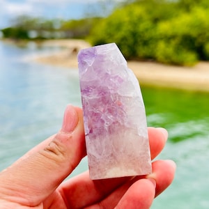 May include: A hand holds a polished amethyst crystal point. The crystal transitions from a pale, translucent base to a vibrant purple hue at the top. The background features a beach, water, and greenery.