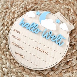 Può includere: Un cartello rotondo in legno per l'annuncio della nascita di un bambino con le parole "hallo Welt" in corsivo azzurro. Il cartello include spazio per scrivere il nome, la data, il peso, l'ora e la taglia del bambino. Decorato con una luna blu, nuvole bianche e stelle.