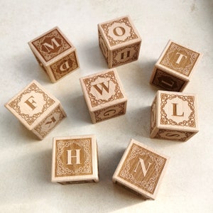 Könnte beinhalten: Acht Holzbuchstabenwürfel mit gravierten Buchstaben und dekorativen Bordüren. Die Würfel sind auf einer hellen Oberfläche angeordnet. Jeder Würfel zeigt einen anderen Buchstaben, darunter M, O, T, W, L, H, N und F.