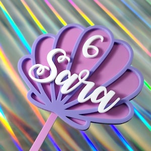 May include: A cake topper in the shape of a seashell, featuring the name "Sara" and the number "6" in white. The shell is purple and pink, with a light purple stem. The background is iridescent.