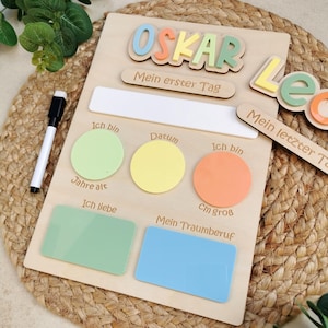May include: A light-coloured wooden milestone board featuring the name OSKAR in pastel 3D letters, with German text labels for age, date, height, interests, and dream job. It includes a white dry-erase section, coloured acrylic inserts, and a black marker.