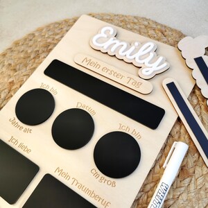 May include: A light-coloured wooden milestone board featuring the name Emily in raised white script. The board includes black magnetic sections for writing details like age, date, height, and dream job, accompanied by a white marker pen.