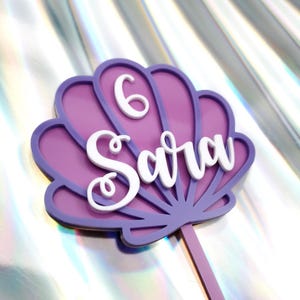 May include: A purple and lavender seashell-shaped cake topper with the name "Sara" in white script and the number "6". The topper is on a lavender stick, ideal for a birthday cake.