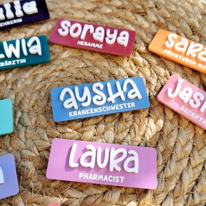 Op de afbeelding: Een verzameling kleurrijke naambadges met witte letters. De badges zijn gemaakt van een dik, duurzaam materiaal en hebben afgeronde hoeken. De namen op de badges zijn "Soraya", "Aysha", "Laura", "Sara", "Jas" en "Lia". Elk badge bevat ook een functietitel, zoals "Hebamme", "Krankenschwester", "Pharmacist", "Registered Nurse", "Receptionist" en "Ehrerin".