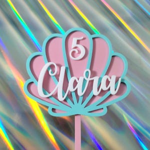 May include: A pink and turquoise seashell cake topper with the name "Clara" in white script and the number "5". The topper is on a pink stick, ideal for a birthday cake.