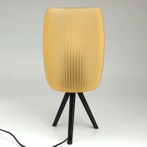 Könnte beinhalten: Moderne Tischlampe mit einem zylindrischen, gerippten, gelben Lampenschirm. Der Lampenschirm steht auf einem schwarzen Dreibein-Fuß. Die Lampe ist mit einem schwarzen Kabel an eine Steckdose angeschlossen. Dekorative Beleuchtung.