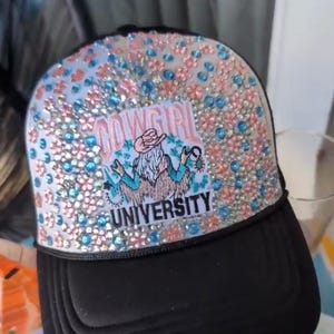 May include: Black trucker hat with a bedazzled top featuring pink and blue rhinestones. The hat has a patch that reads "Cowgirl University" with a graphic of a cowgirl. The hat is a stylish accessory.