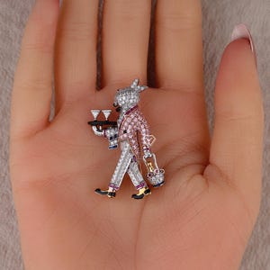 以下が含まれることがあります： ピンクと白のジャケットを着たウサギのウェイターの形をしたシルバーのブローチ。2つのグラスとバスケットが入ったトレイを持っています。ブローチは透明とピンクのクリスタルで装飾され、黒と金のアクセントが付いています。