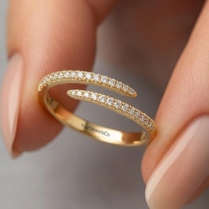 May include: A gold-colored ring with a spiral design, encrusted with small, sparkling gemstones. The ring is held between two fingers, showcasing its elegant and delicate craftsmanship. The text "YuviJewelsCo" is visible on the ring.