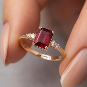 May include: A gold ring with a rectangular, deep red gemstone. The ring is set with smaller, clear gemstones on either side of the main stone. The band is slender and polished, with the text "YuviJewelsCo" inscribed on the inside.