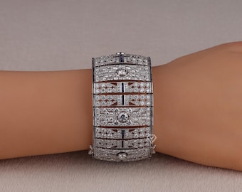 May include: A silver-toned bracelet with intricate detailing, featuring numerous small, sparkling stones and a geometric design. The bracelet has a rectangular shape and is composed of several connected segments. Small blue stones are interspersed throughout the design.